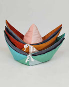 Vier farbenfrohe, handgefertigte, dreieckige, weiche Aufbewahrungskörbe mit Kordelzug sind in einem Farbverlauf von Blaugrün unten nach Orange und Rosa oben gestapelt und werden vor einem schlichten Hintergrund präsentiert. Der Stapel erinnert an die verspielten Designelemente der Spielmatte und Krabbeldecke Boot von Main Design.
