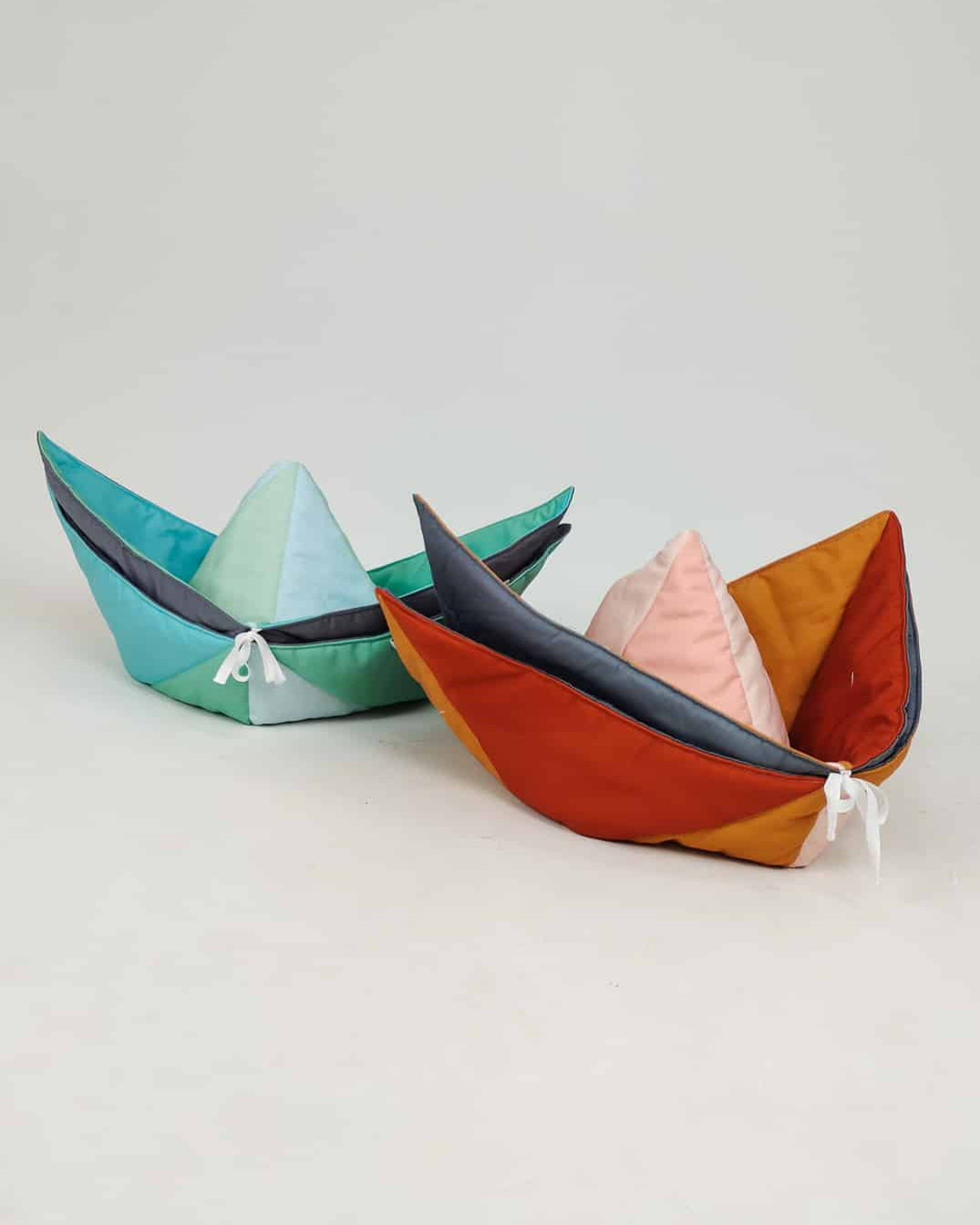 Zwei plüschige Kissen in Papierbootform aus der Kollektion Spielmatte und Krabbeldecke Boot von Main Design liegen verspielt auf dem Boden. Ein Kissen ist in den Farben Blaugrün und Marineblau gehalten, während das andere Pfirsich-, Schwarz- und Rosttöne kombiniert. Beide sind vorne mit zarten weißen Kordeldetails verziert.