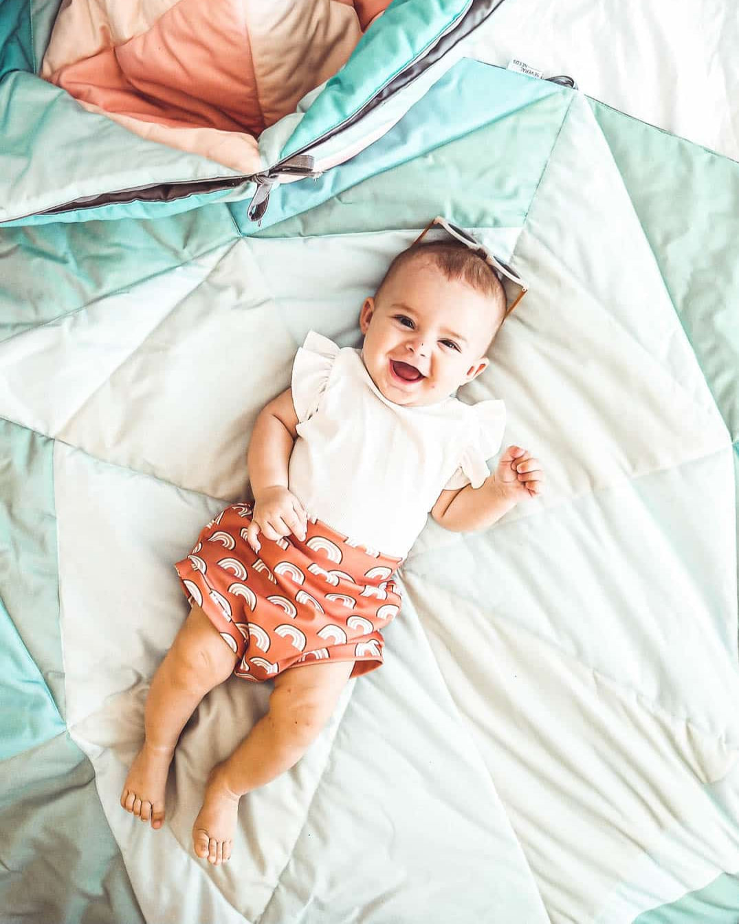 Ein lächelndes Baby in einem weißen Shirt mit Regenbogenmuster und orangefarbenen Shorts liegt auf der Spielmatte und Krabbeldecke Boot von Main Design und trägt eine Sonnenbrille auf dem Kopf.