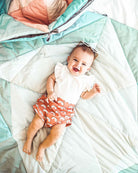 Ein lächelndes Baby in einem weißen Shirt mit Regenbogenmuster und orangefarbenen Shorts liegt auf der Spielmatte und Krabbeldecke Boot von Main Design und trägt eine Sonnenbrille auf dem Kopf.