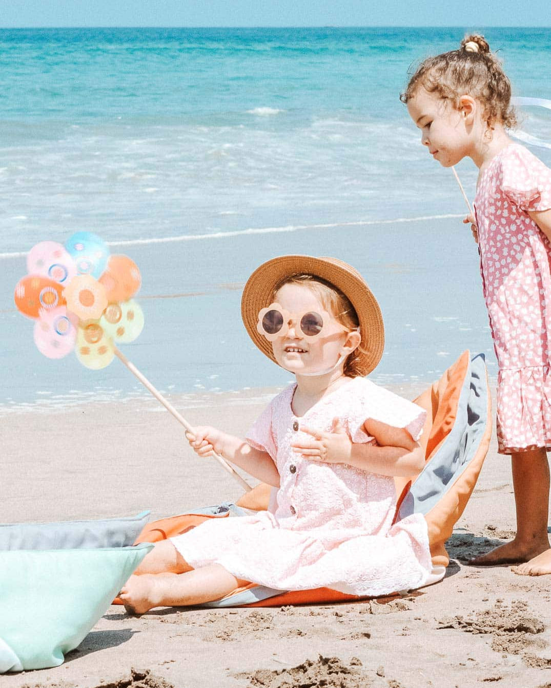 Zwei kleine Kinder genießen den Tag an einem Sandstrand. Eines sitzt gemütlich auf der Spielmatte und Krabbeldecke Boot von Main Design, hält ein handgefertigtes Windrad in der Hand und trägt eine Sonnenbrille und einen Hut. Das andere steht in einem rosa Kleid gekleidet und staunt über das Windrad. Im Hintergrund rauschen sanft die Meereswellen.