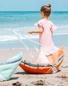 Ein junges Mädchen mit Duttfrisur in einem rosa-weißen Kleid hält eine Drachenschnur und steht am Sandstrand, wo zwei bunte Drachen in der Nähe ruhen. Die Meereswellen und der klare blaue Himmel rahmen die Szene ein und erinnern an eine luftige Main Design Spielmatte und Krabbeldecke Boot.