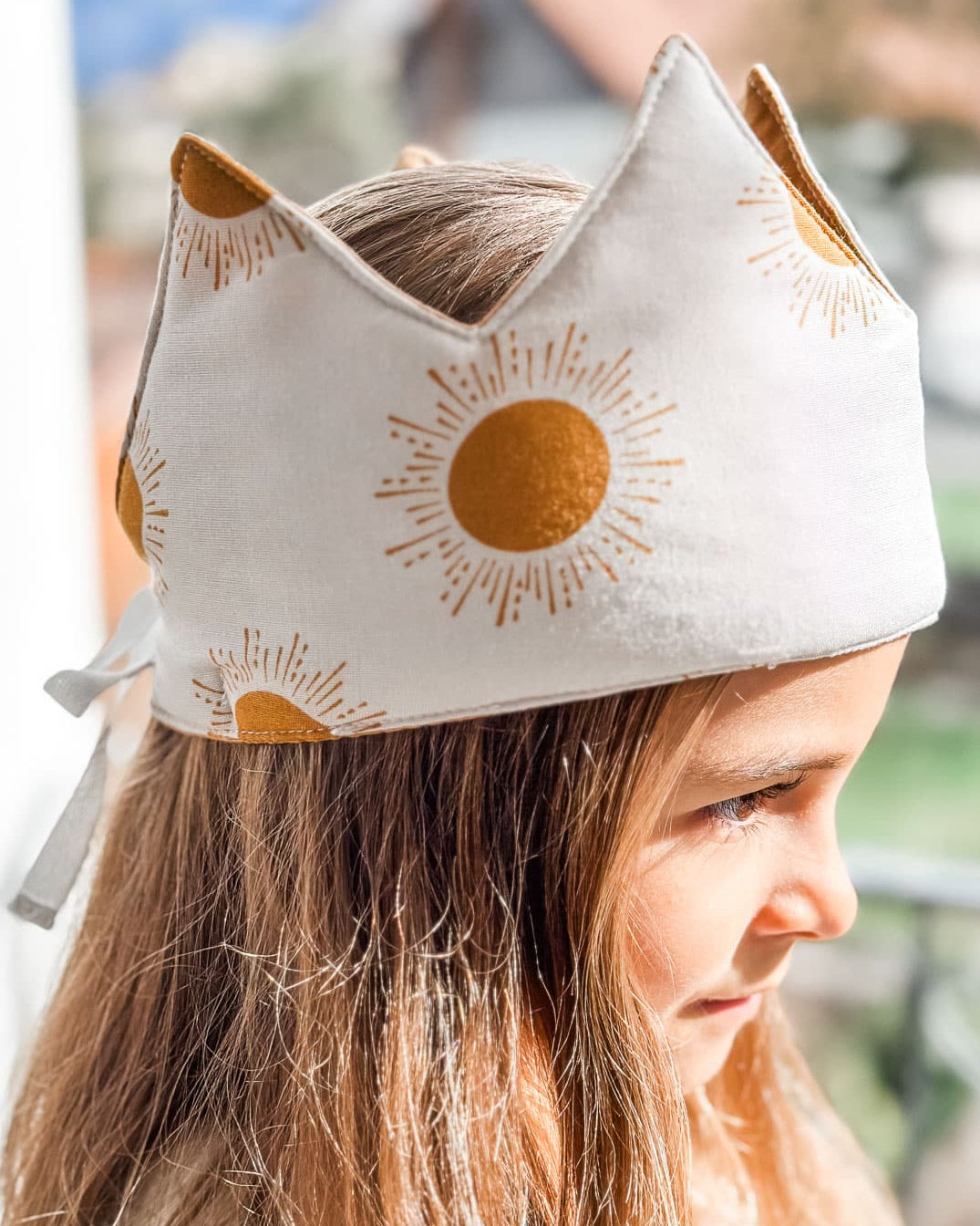 Ein kleines Kind mit hellbraunem Haar lächelt und schaut im natürlichen Sonnenlicht zur Seite. Es trägt eine wendbare Stoffkrone von Several Needs by Main Design, die mit goldenen Sonnenmustern verziert ist.