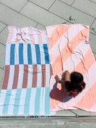 Ein Kind mit langen dunklen Haaren sitzt auf einem rosa und orange gestreiften Main Design Strandtuch Serra aus recycelter Baumwolle, daneben ein blau, rosa und braun gestreiftes Handtuch, beide ausgebreitet auf hellem Pflaster.