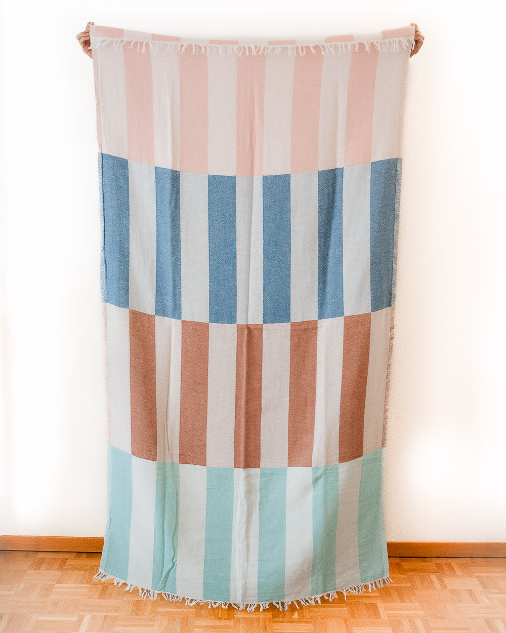 Das Strandtuch Blockberry von Main Design ist rechteckig, mit kurzen Fransen und vertikalen Pastellstreifen in Pink, Blau, Braun und Mintgrün - erinnert an ein buntes Strandtuch, flach an einer weißen Wand über Holzboden hängend.