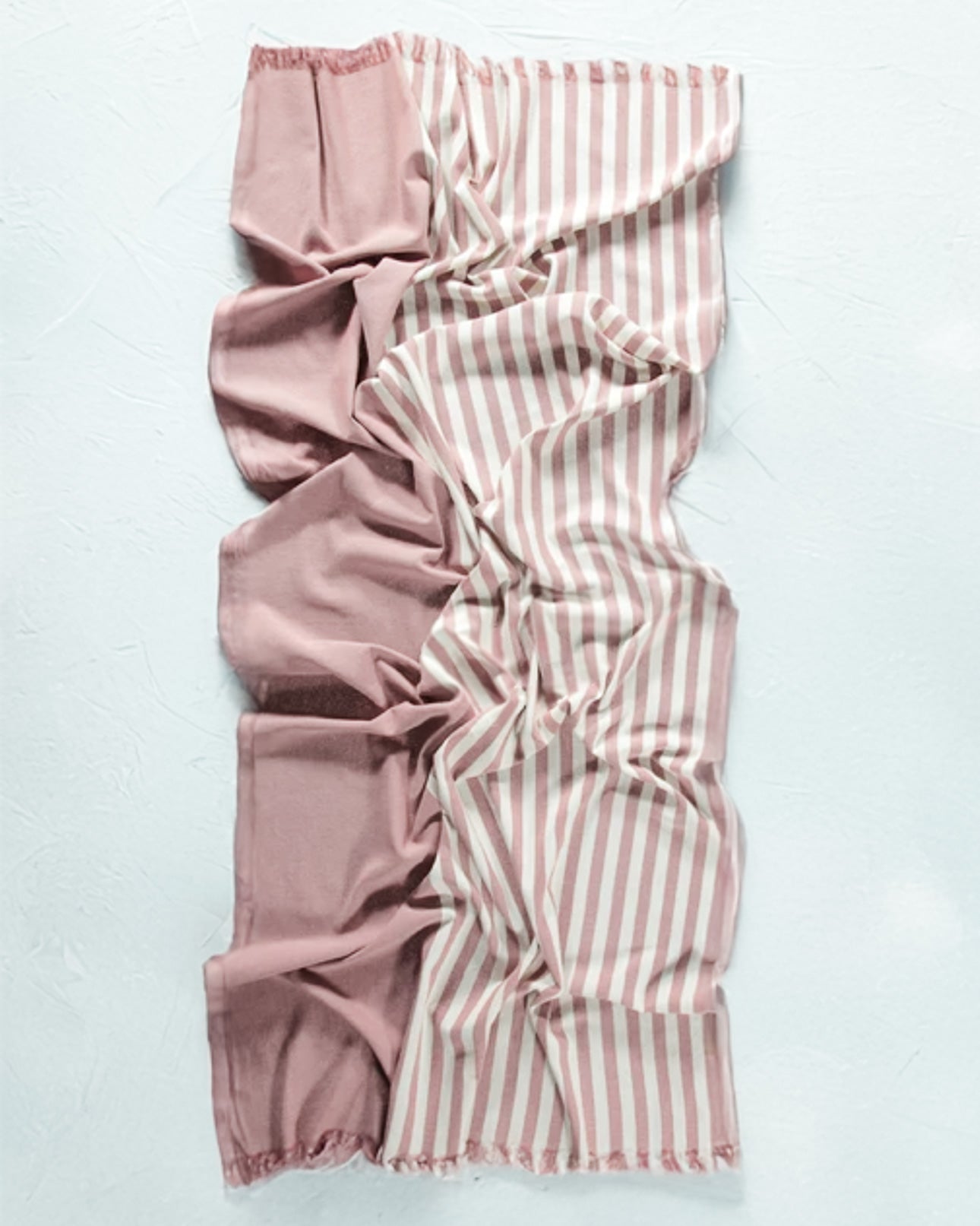 Ein zerknittertes rechteckiges Strandtuch Fiesta von Main Design, halb einfarbig staubrosa, halb rosa und weiß gestreift, auf einer hellen Oberfläche. Hergestellt aus Baumwolle und OEKO-TEX-zertifiziert für Qualität und Sicherheit.