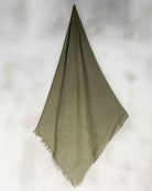 Ein einzelnes olivgrünes Main Design Strandtuch Nomad aus OEKO-TEX zertifizierter Baumwolle mit Fransen hängt diagonal vor einem hellen, verschwommenen Hintergrund.