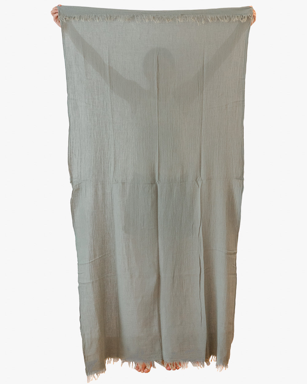Eine Person hält das Strandtuch Nomad von Main Design hoch, ein großes, hellgrau-grünes, strukturiertes Handtuch mit ausgefransten Rändern aus OEKO-TEX-zertifizierter Baumwolle, das ihren Körper bis auf Finger und Zehen verdeckt.