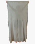 Eine Person hält das Strandtuch Nomad von Main Design hoch, ein großes, hellgrau-grünes, strukturiertes Handtuch mit ausgefransten Rändern aus OEKO-TEX-zertifizierter Baumwolle, das ihren Körper bis auf Finger und Zehen verdeckt.