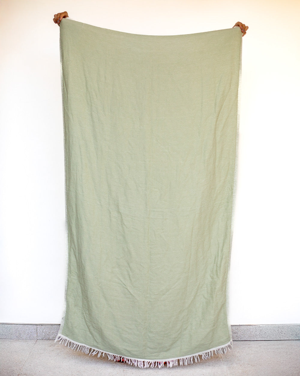 Eine Person steht vor einer weißen Wand und hält das große, hellgrüne Strandtuch Zen von Main Design hoch. Das Handtuch aus OEKO-TEX-Baumwolle hat ausgefranste Ränder, hinter dem strandtauglichen Stoff sind nur ihre Hände und Schuhspitzen zu sehen.