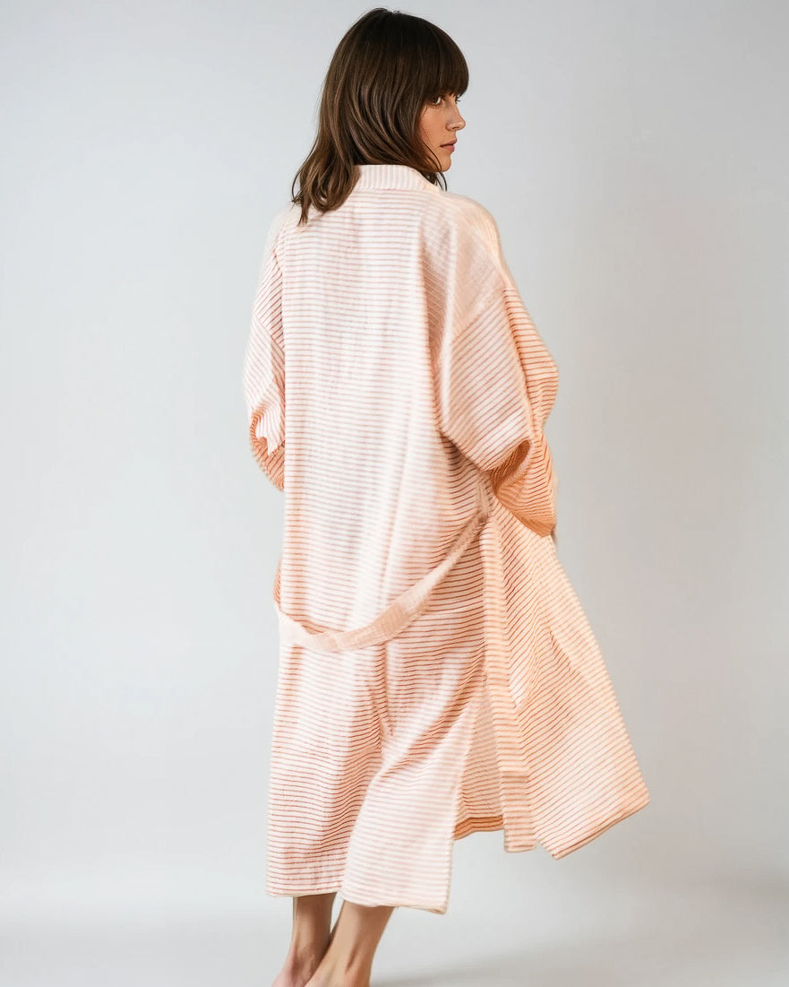 Eine barfuß gehende Frau mit schulterlangen braunen Haaren steht mit dem Rücken zur Wand und trägt den Kimono Sunset Stripes von Main Design mit hellrosa und weißen Streifen vor einem unifarbenen Hintergrund.