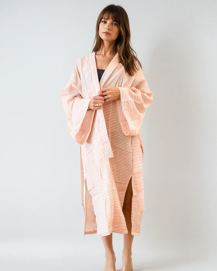Eine Frau steht vor einem unifarbenen Hintergrund und trägt den Sunset Stripes Kimono von Main Design, ein langes, lockeres pfirsich-weiß gestreiftes Gewand mit weiten Ärmeln aus gekämmter Baumwolle, das über ein dunkles Oberteil für entspannte Strandmode gelegt wird.