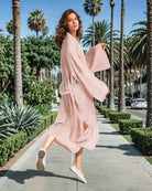 Eine Frau im Main Design Sunset Stripes Kimono und weißen Slippern springt spielerisch auf einem sonnigen, von Palmen und Grün gesäumten Bürgersteig.