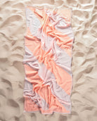 Das Strandtuch Serra von Main Design, ein pastellfarbenes, pfirsichfarbenes und weißes Handtuch aus recycelter Baumwolle mit geometrischem Muster, liegt leicht zerknittert auf einem Sandstrand und ist von strukturiertem Sand umgeben.