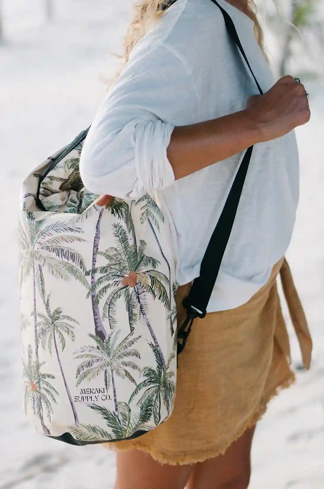 Eine Person in einem weißen Hemd und hellbraunen Shorts trägt einen Dry Bag von Meraki Supply Co. mit tropischem Motiv, der mit einem Palmenmuster und dem Text „Meraki Supply Co.“ von Main Design verziert ist. Der Hintergrund scheint ein Sandstrand zu sein.