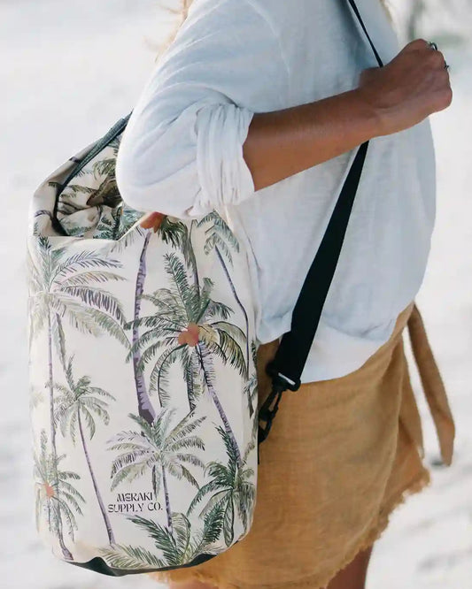 Eine Person in einem weißen Hemd und hellbraunen Shorts trägt einen Dry Bag von Meraki Supply Co. mit tropischem Motiv, der mit einem Palmenmuster und dem Text „Meraki Supply Co.“ von Main Design verziert ist. Der Hintergrund scheint ein Sandstrand zu sein.