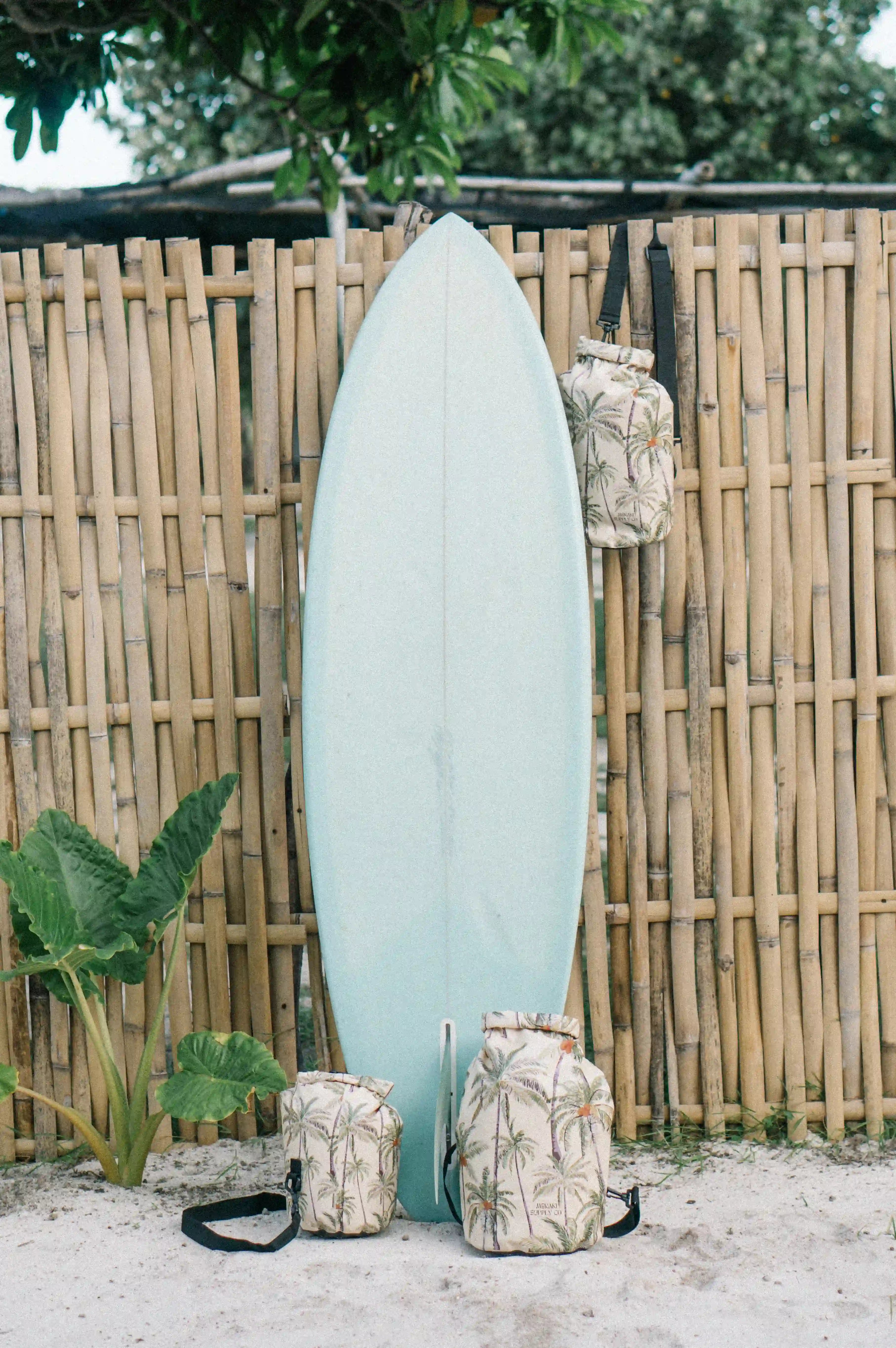 Ein hellblaues Surfbrett lehnt an einem Bambuszaun. Drei Main Design Dry Bag von Meraki Supply Co.-Taschen mit Palmenmotiven hängen am Zaun und liegen im Sand. Im Hintergrund sind üppige grüne Pflanzen und Bäume zu sehen.