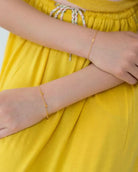 Eine Person in einem leuchtend gelben Kleid sitzt mit verschränkten Armen da und trägt zwei zarte Armbänder von Adorabili an ihren Handgelenken.