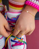 Ein Kind bindet regenbogenfarbene Schnürsenkel an weiße Turnschuhe, trägt bunt gestreifte Socken, leuchtend rosa Perlenarmbänder und ein zartes handgefertigtes Armband von Adorabili by Adorabili.