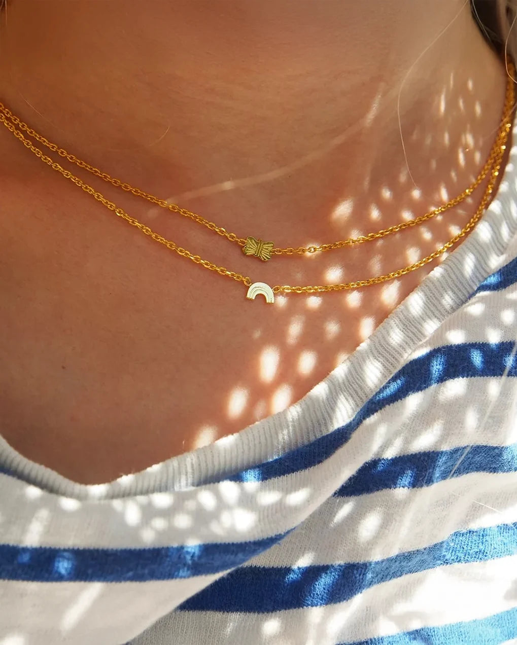 Nahaufnahme der Halskette von Adorabili: Zwei 24K-vergoldete Ketten, eine mit Palmblatt-Anhänger, eine mit Regenbogen, getragen über einem blau-weiß gestreiften Shirt im Sonnenlicht. Perfektes Geschenk für Kinder.