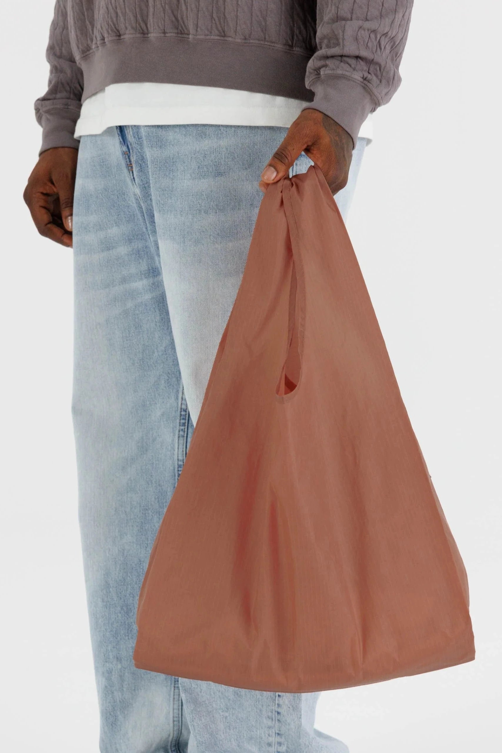 Eine Person mit dunklem Hautton hält eine braune BAGGU Standard Tragetasche von Main Design. Sie trägt ein graues Langarmshirt und hellblaue Jeans vor einem schlichten weißen Hintergrund.