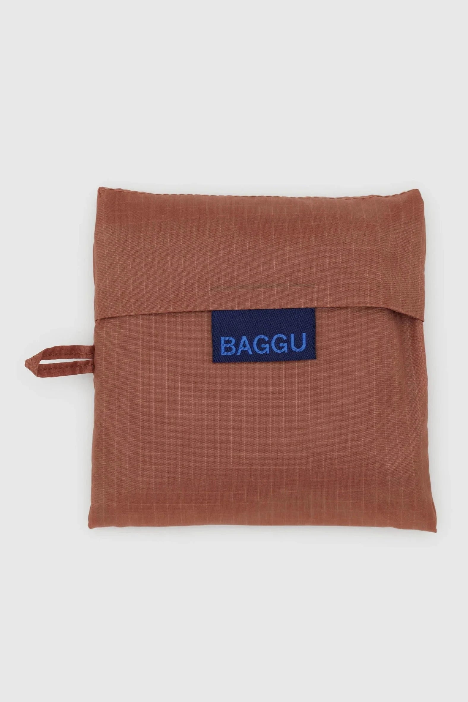 Eine kompakte, gefaltete, rostfarbene Mehrwegtasche der Marke Main Design. Die BAGGU Standard Tragetasche aus recyceltem Ripstop-Nylon hat eine ordentliche quadratische Form und eine Klappe, die durch ein aufgenähtes Etikett mit dem Wort „BAGGU“ in blauer Schrift gesichert ist. Auf der linken Seite ist eine kleine Schlaufe zum Aufhängen oder Tragen sichtbar.