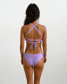 Eine Frau blickt von der Kamera weg und trägt das Charm Surf Bikini Oberteil von Main Design in Hellviolett mit überkreuzten Rückenträgern aus chlorresistentem Stoff – ideal für Wassersportarten, vor einem schlichten weißen Hintergrund.