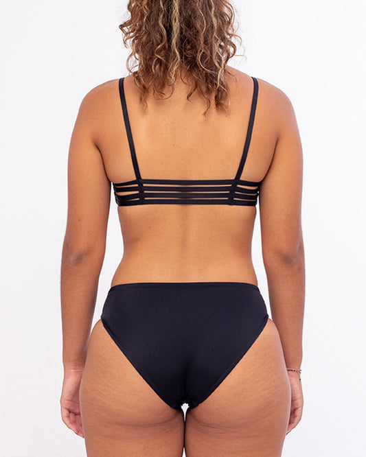 Eine Person mit lockigem Haar steht mit dem Rücken zur Kamera und präsentiert die schwarze Bliss Bikinihose von Main Design mit Riemchendetails am Oberteil und hoch tailliertem Design. Dieser nachhaltige Bikini aus umweltfreundlichem Stoff bietet einen auffälligen Kontrast zum schlichten, hellen Hintergrund.