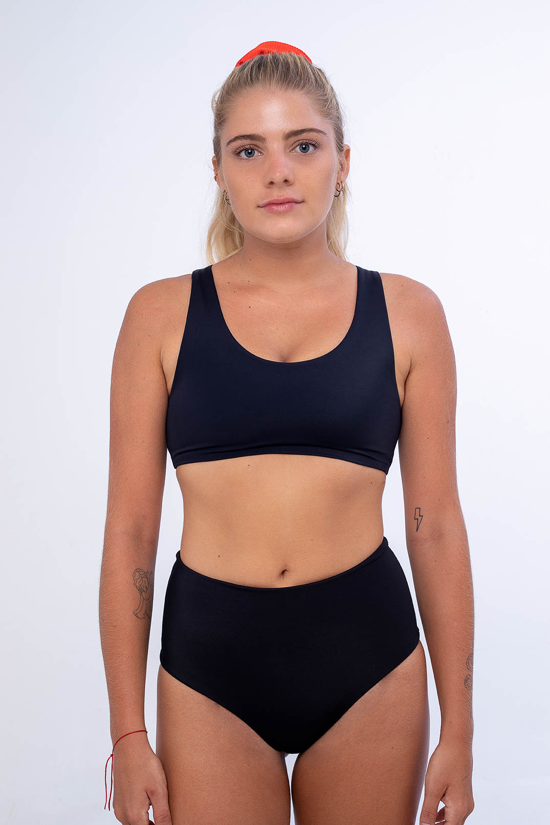 Vor einem schlichten weißen Hintergrund trägt eine Person mit zurückgebundenem langen Haar einen schwarzen Sport-BH aus recycelten Materialien, kombiniert mit der Posh Bikini Hose von Main Design – einem nahtlosen Unterteil mit hoher Taille. Sie zeigt kleine Tattoos auf jedem Arm und behält einen neutralen Ausdruck bei.