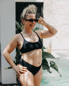 Eine Frau mit blondem Haar im Dutt, Sonnenbrille und schwarzem Bikini mit Ember Bikini Hose von Main Design steht neben einem Pool. Sie lächelt, hat eine Hand auf der Hüfte und rückt mit der anderen ihre Sonnenbrille zurecht. Im Hintergrund sind große grüne Blätter und eine Steinmauer zu sehen.