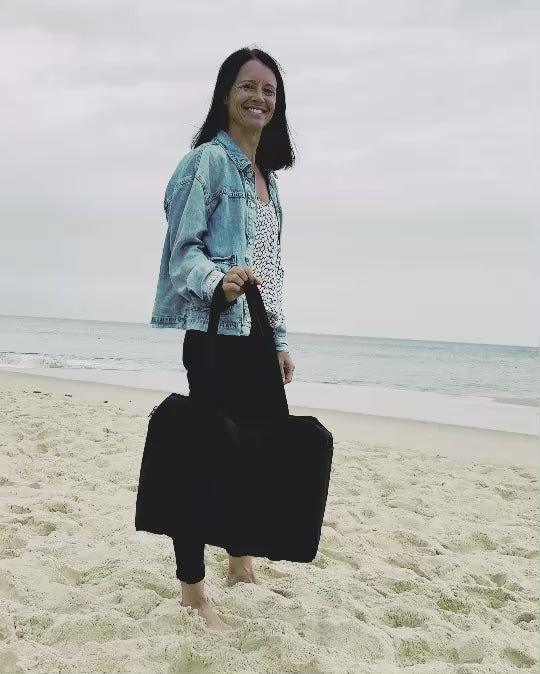 Eine Person mit langen dunklen Haaren lächelt an einem Strand und hält die elegante schwarze BAGGU Small Cloud Carry-On Tasche von Main Design in der Hand. Sie trägt eine helle Jeansjacke, ein gemustertes Hemd und eine schwarze Hose. Im Hintergrund erstreckt sich der Sandstrand vor dem Meer unter einem bewölkten Himmel.