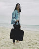 Eine Person mit langen dunklen Haaren lächelt an einem Strand und hält die elegante schwarze BAGGU Small Cloud Carry-On Tasche von Main Design in der Hand. Sie trägt eine helle Jeansjacke, ein gemustertes Hemd und eine schwarze Hose. Im Hintergrund erstreckt sich der Sandstrand vor dem Meer unter einem bewölkten Himmel.